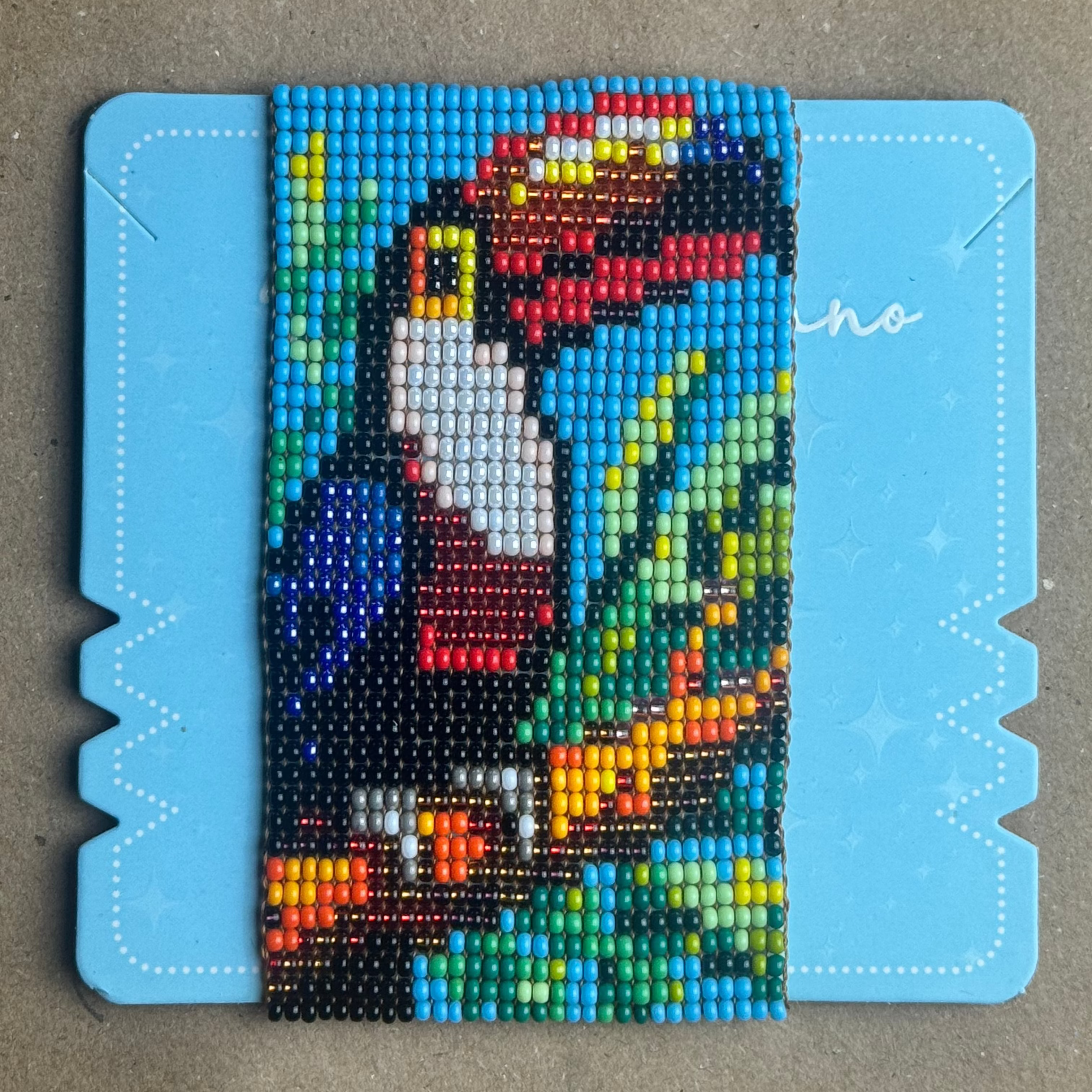 A colorful beaded portrait artwork mounted on a light blue display board, photographed from above on a neutral gray surface.