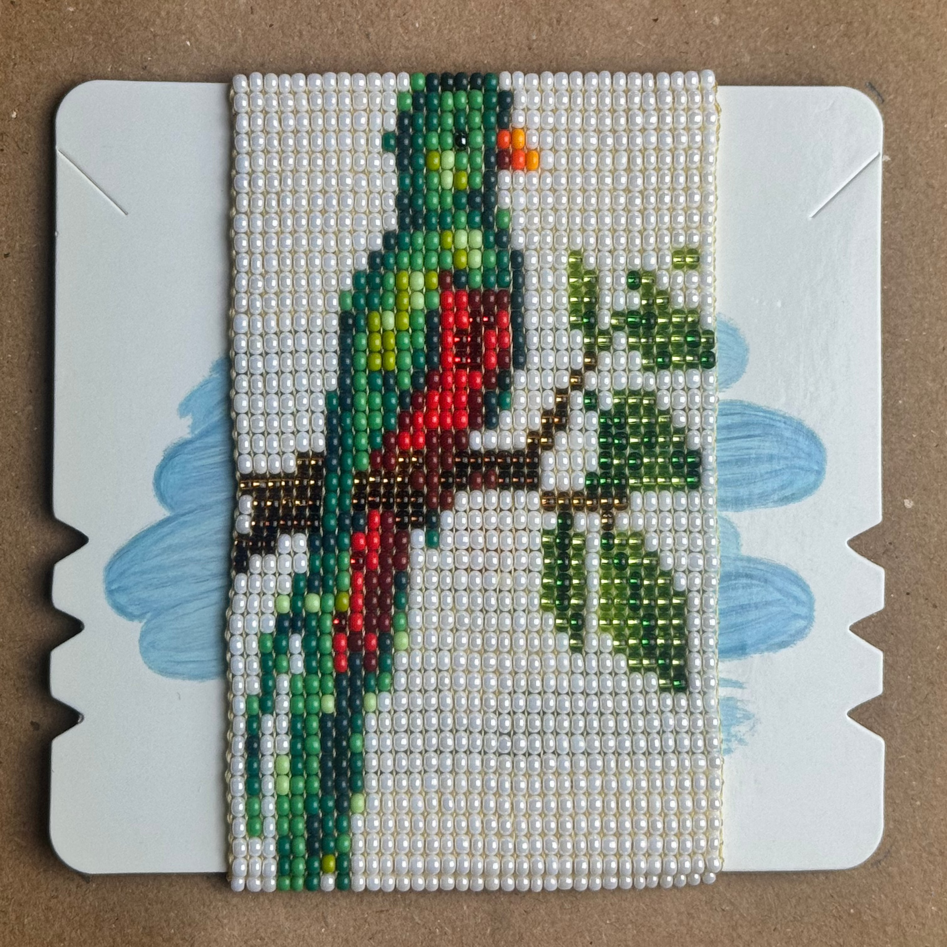 A square beaded craft card featuring a colorful beaded parrot perched on a branch with green leaves, set against a light blue painted background. The card is placed on a brown cardboard surface.