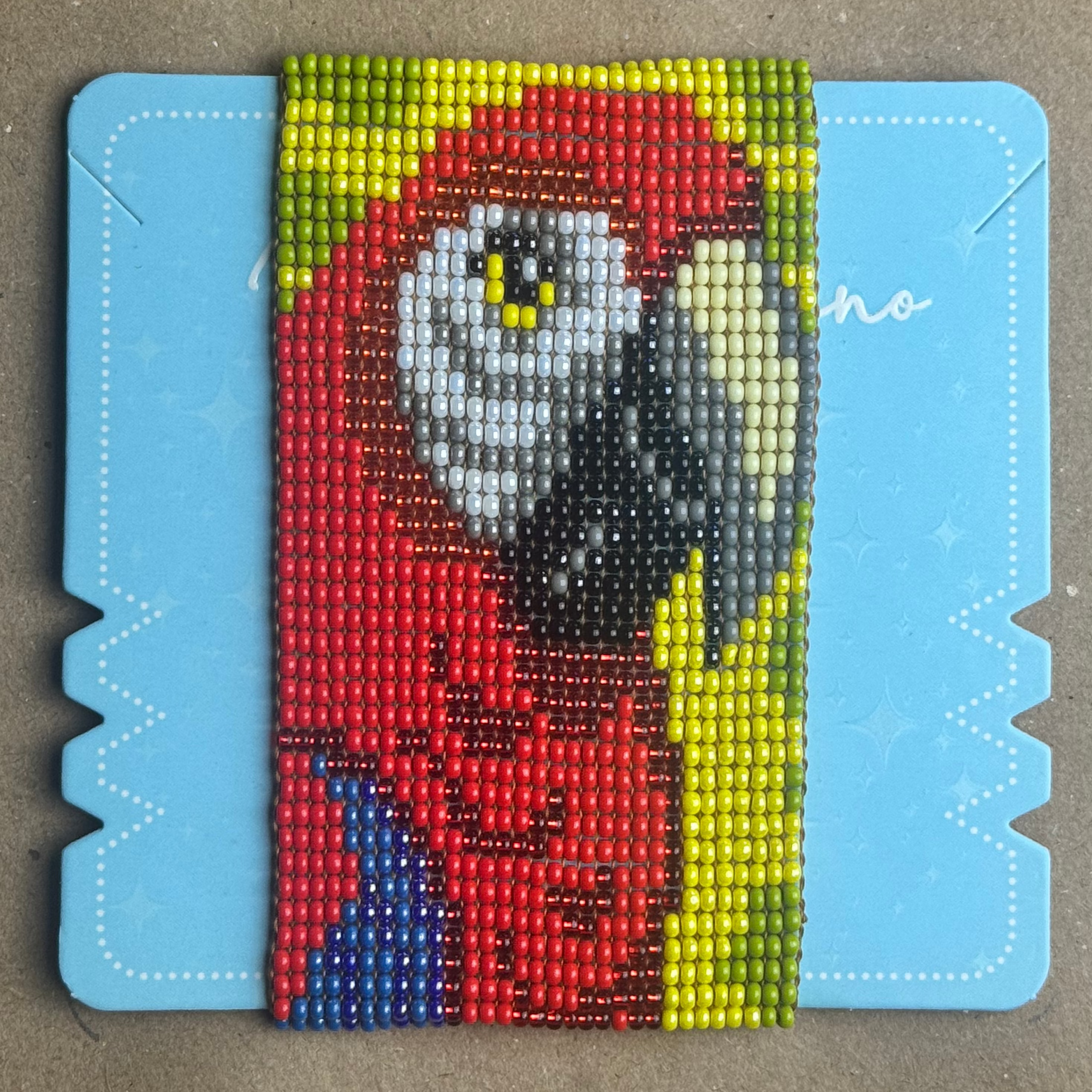A colorful beaded canvas art piece featuring a cartoon character, mounted on a light blue backing board and placed on a brown cardboard background.