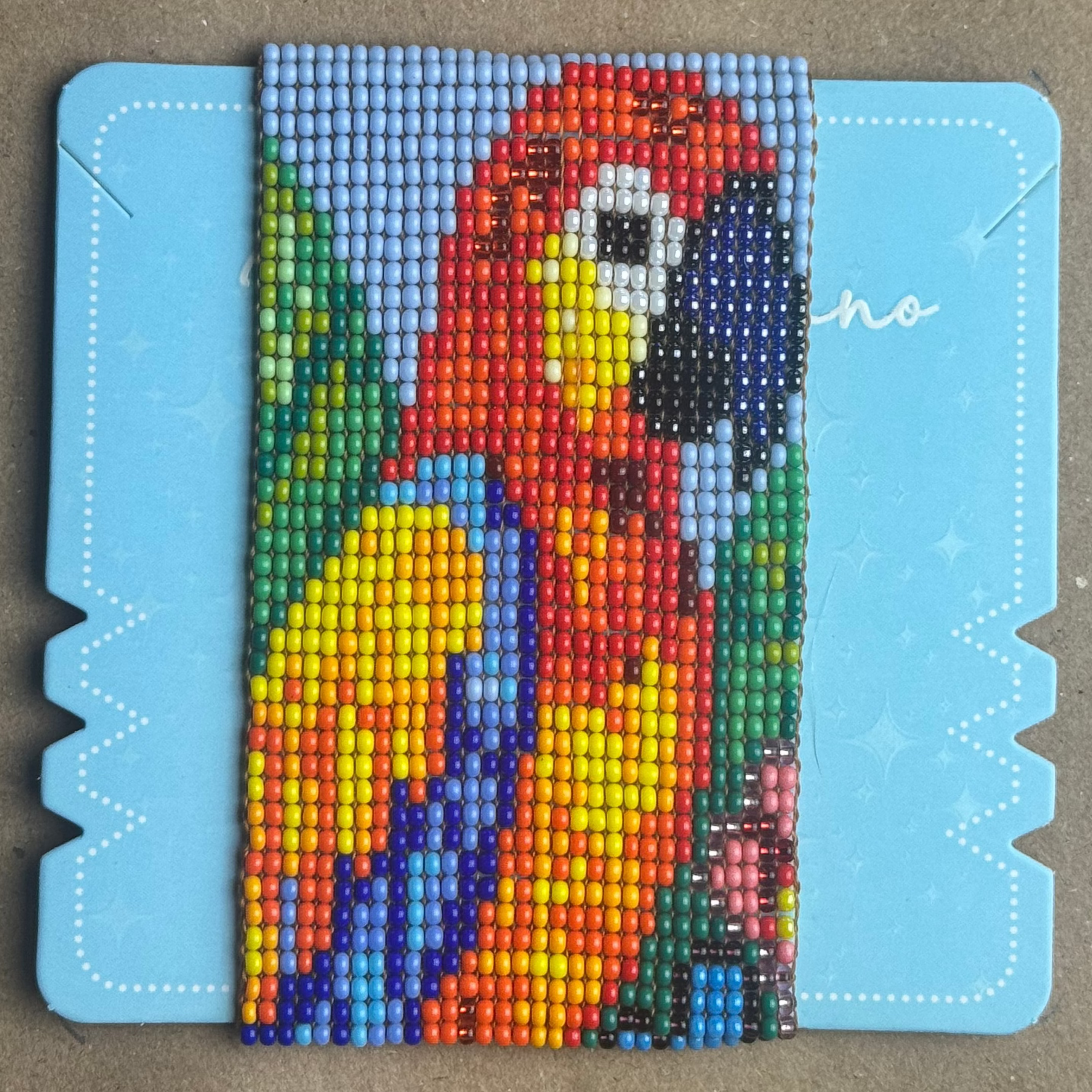 A colorful beaded mosaic panel depicting a parrot, mounted on a light blue plastic frame, placed on a brown cardboard background.