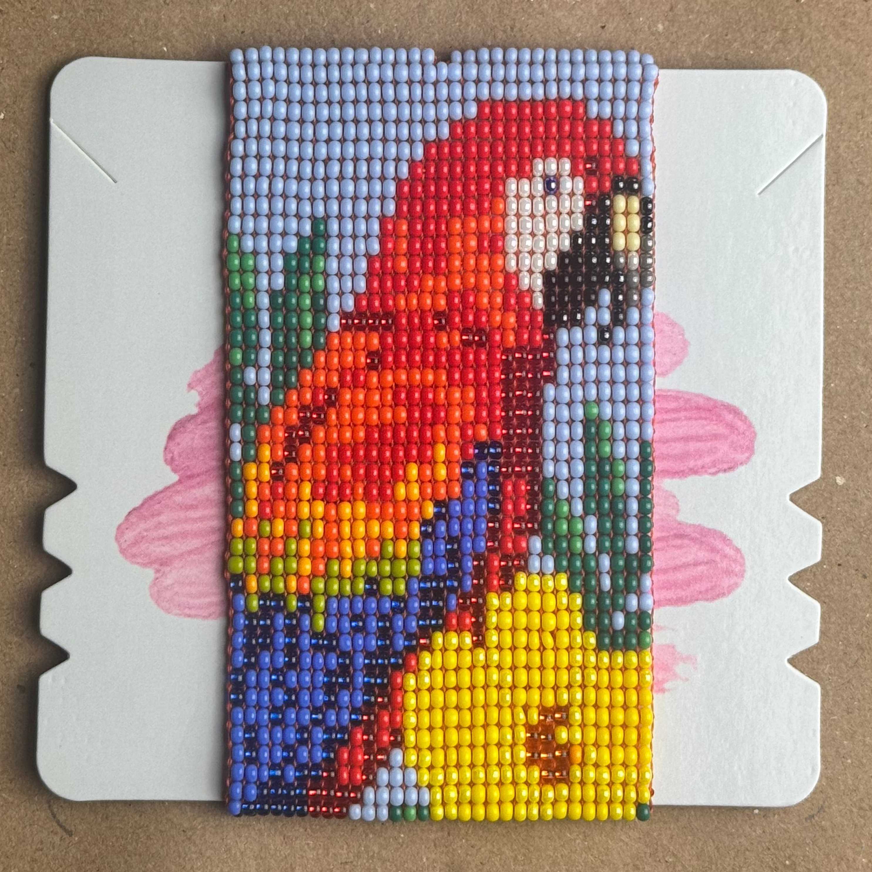 A colorful beaded card featuring a parrot design, displayed on a brown cardboard background.