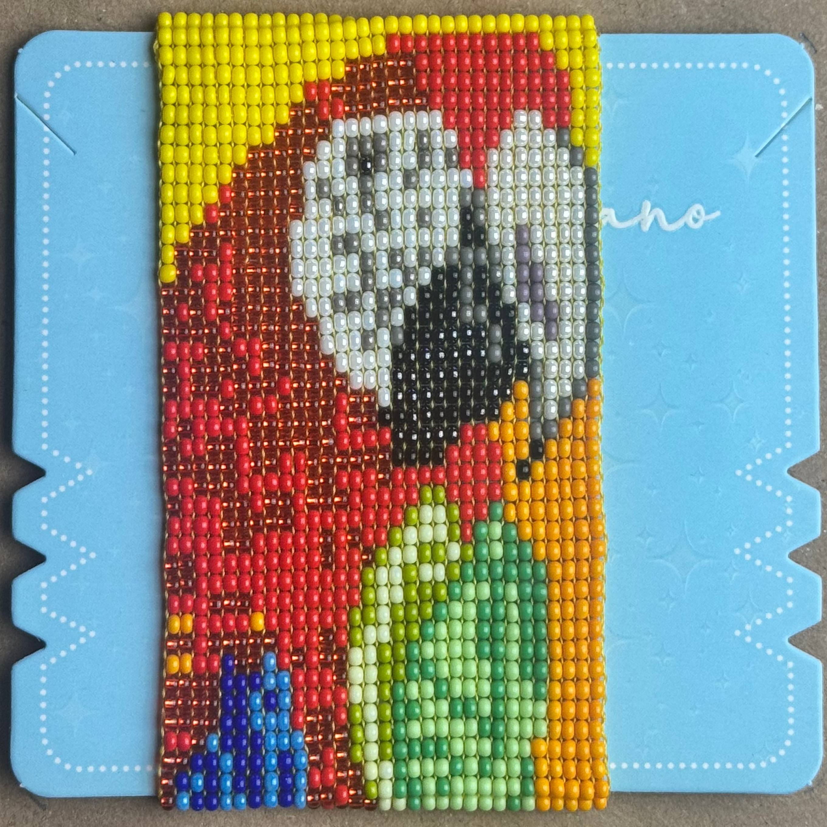 A colorful beaded mosaic art piece depicting a parrot, mounted on a light blue backing board and placed on a brown cardboard background.