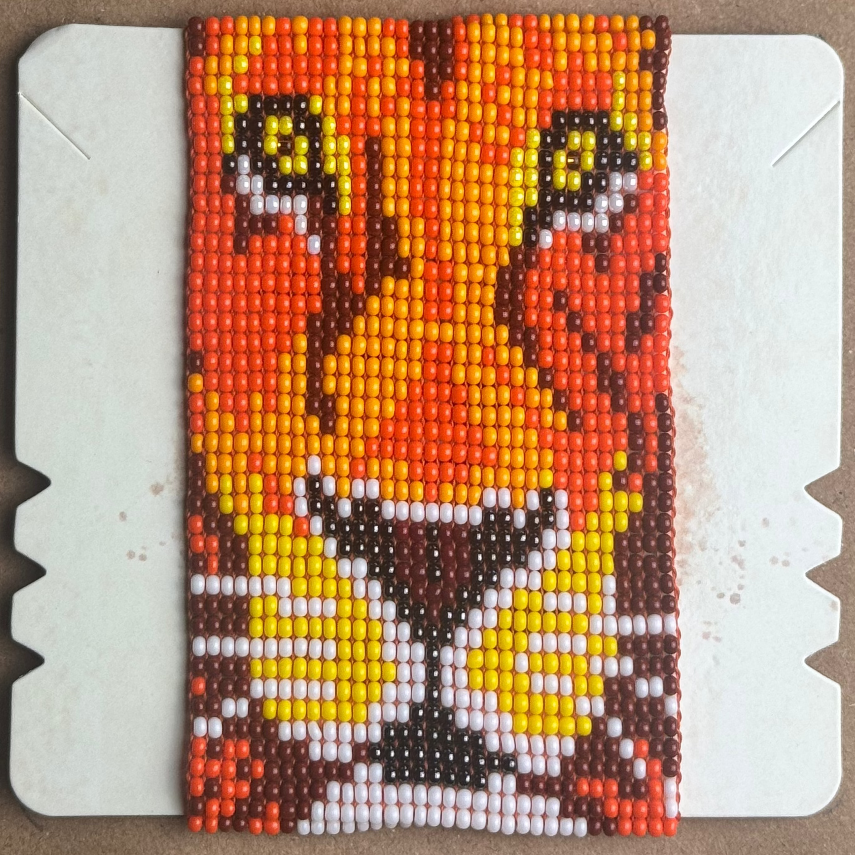 A rectangular beaded patch featuring a stylized cat face in orange, yellow, white, and dark brown, mounted on a white backing card over a brown cardboard background.