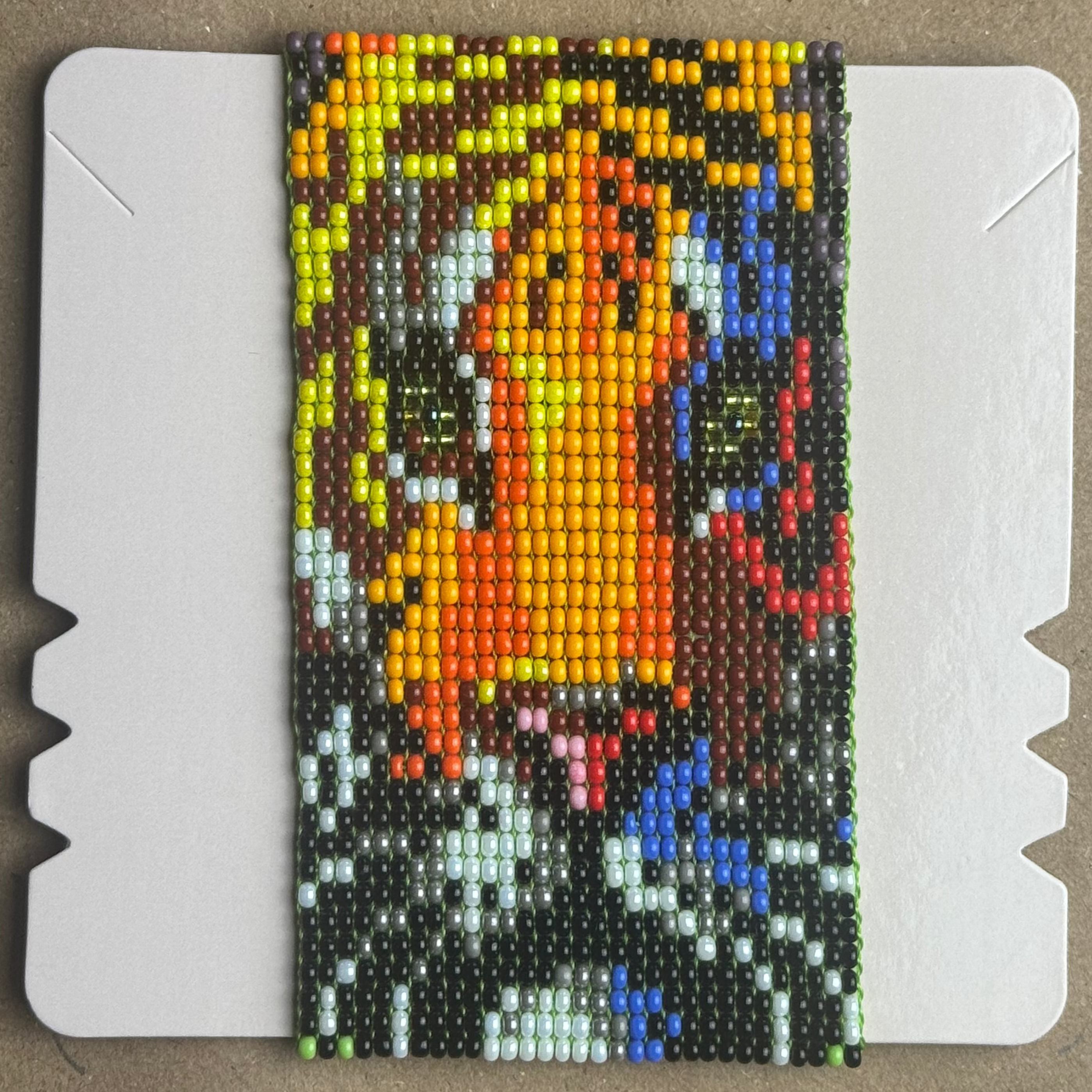 A rectangular beaded card featuring a colorful card-art design, displayed on a white backing board over a brown cardboard surface.