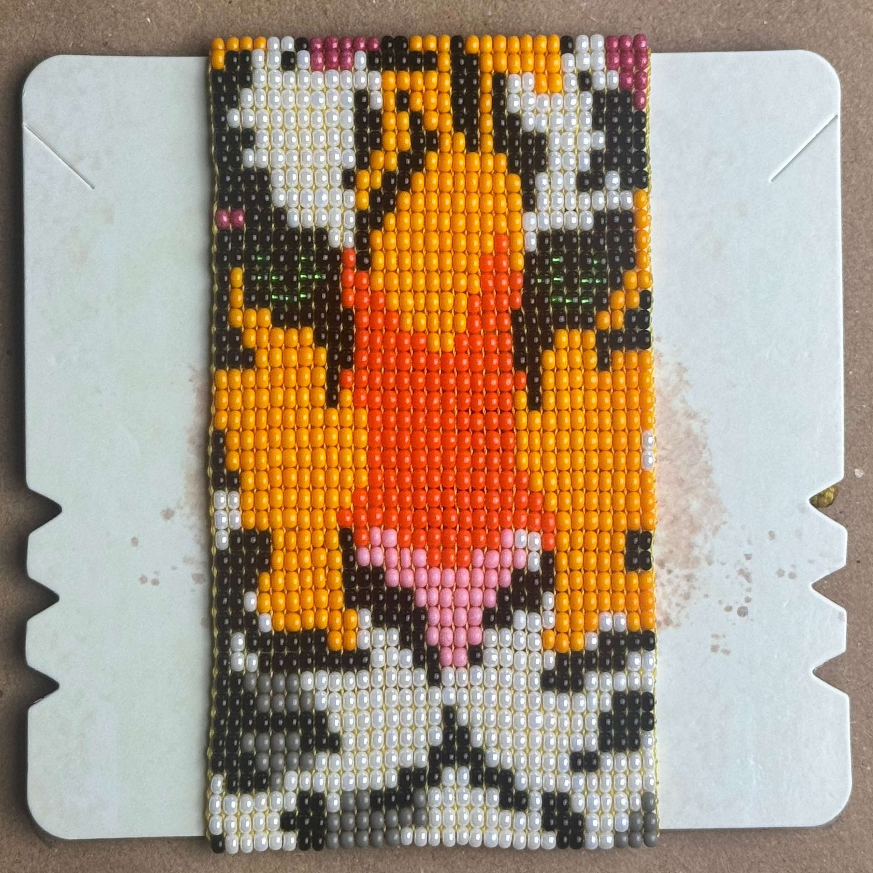 A rectangular beaded textile panel with a symmetrical geometric tiger, mounted on a white plastic display board over a brown cardboard background.