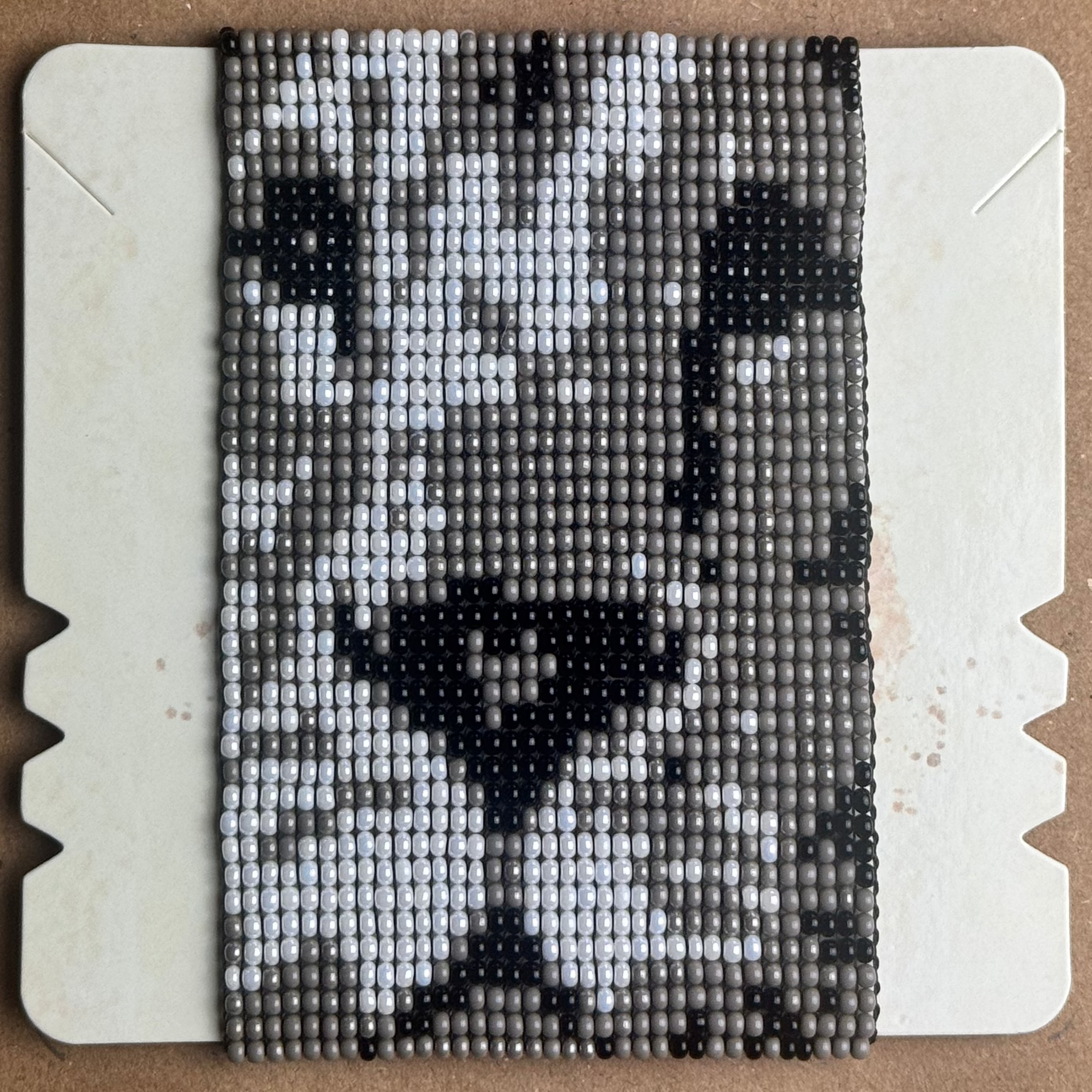 A rectangular beaded textile or mat with a black, white, and gray abstract Lion pattern, displayed flat on a light-colored backing board over a brown cardboard background.