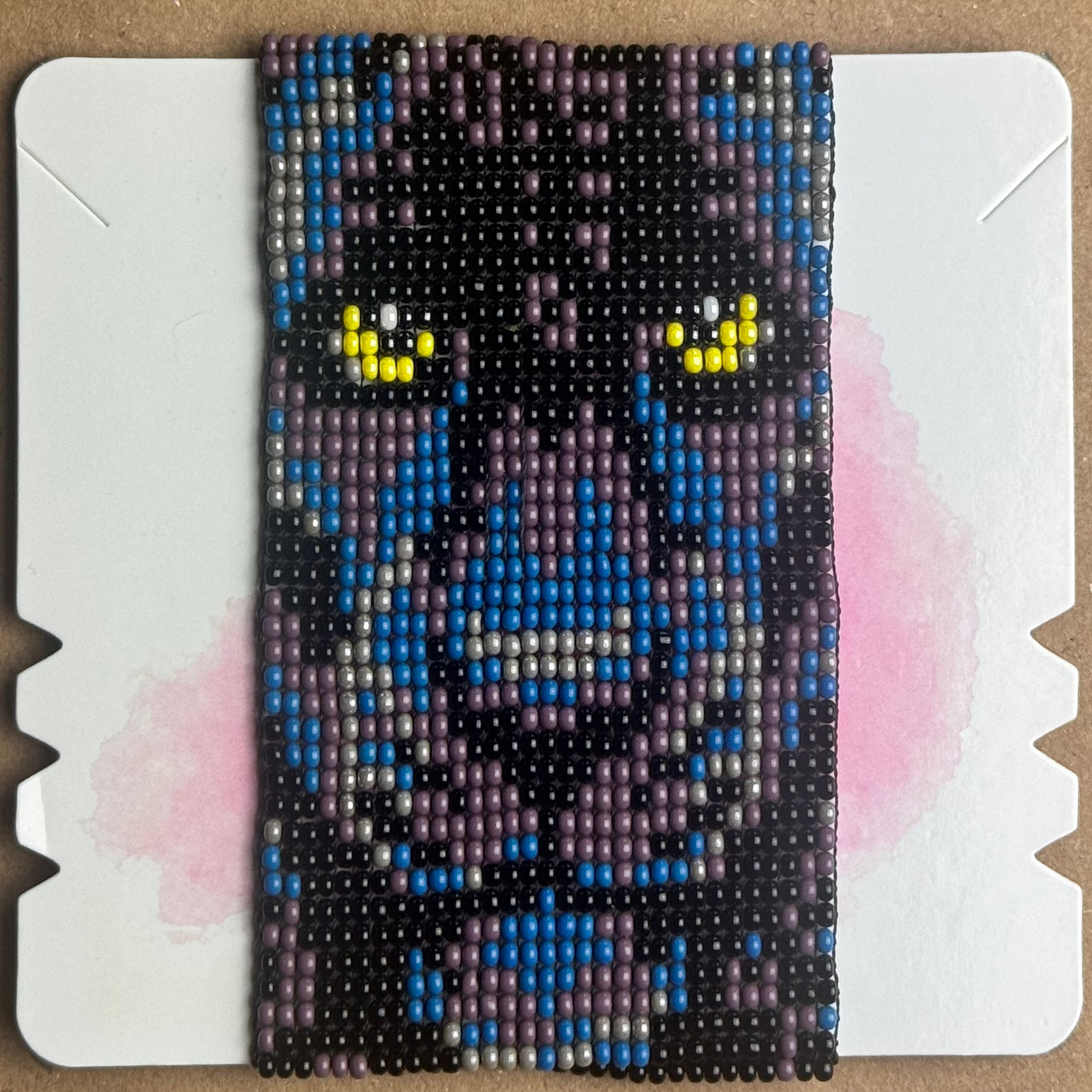 A handmade beaded headband featuring a pixelated puma design in blue, purple, black, and yellow, displayed on a white cardboard backing over a brown kraft paper background.