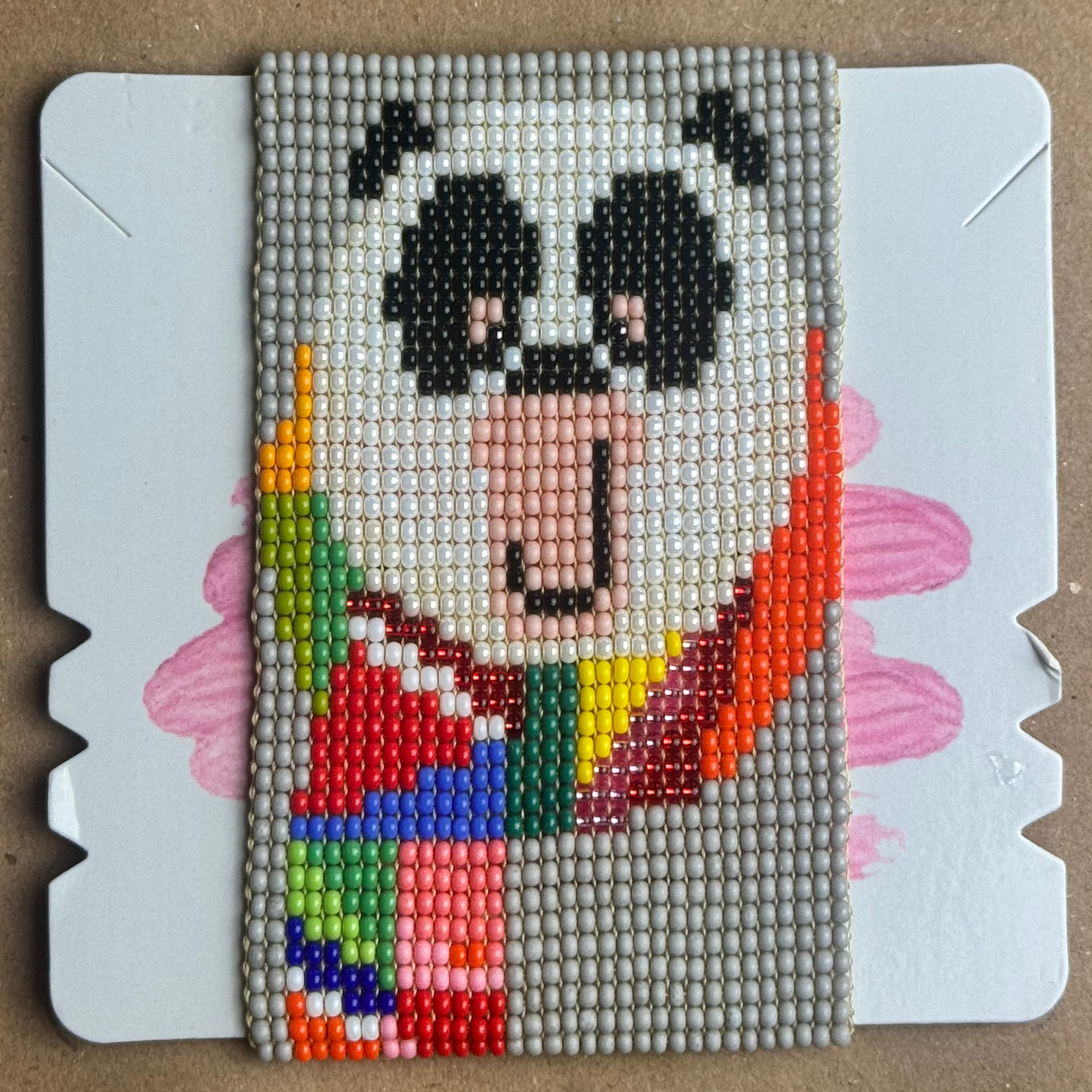 A handmade beaded canvas art piece featuring a stylized panda with a rainbow-colored hair, displayed on a white backing card over a brown cardboard background.