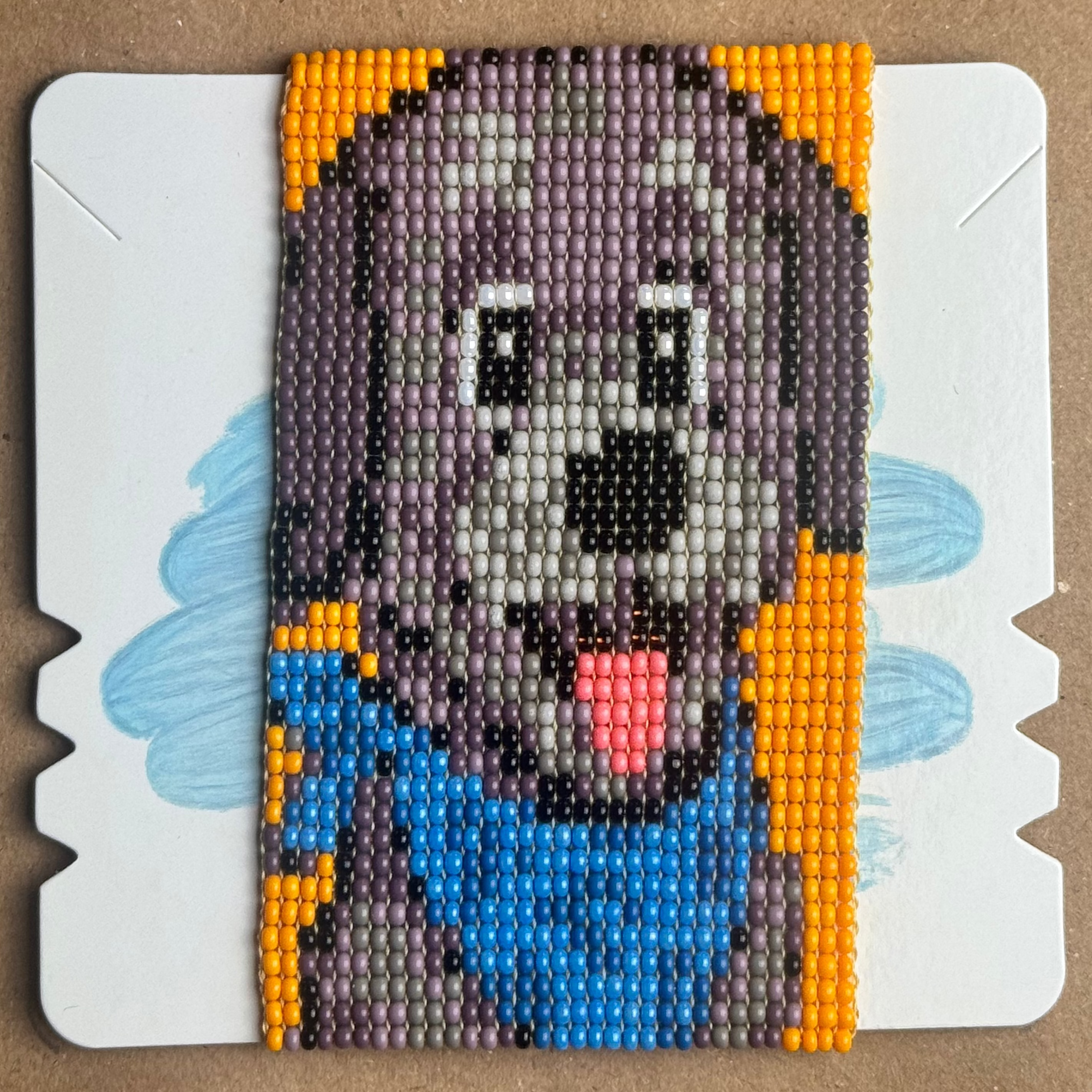 A square beaded art piece featuring a stylized cartoon dog face with a blue hat, gray body, black nose, and orange accents, mounted on a white backing card over a brown cardboard background.