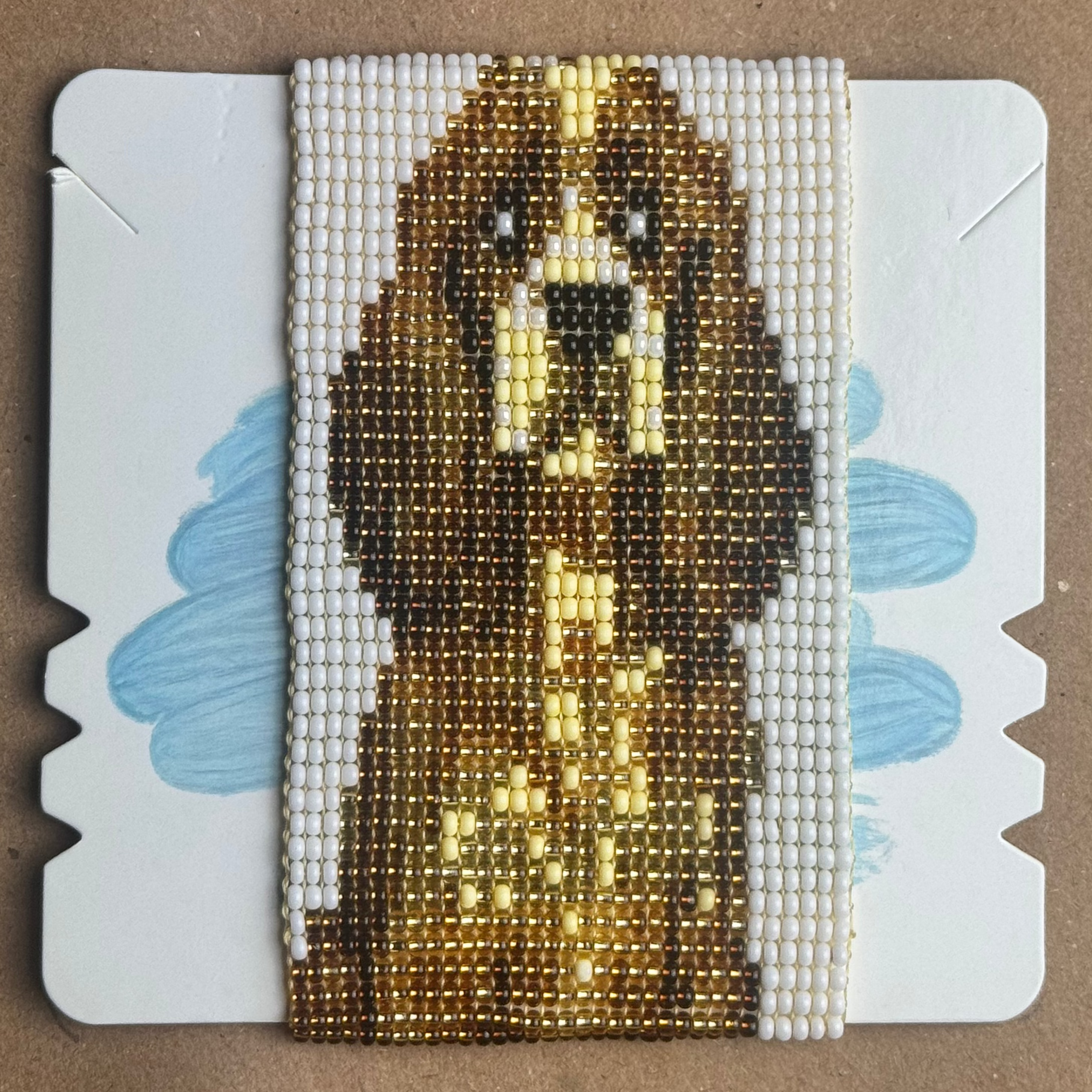A handmade beaded bracelet featuring a brown dog face design, displayed on a white card with blue painted accents against a brown cardboard background.