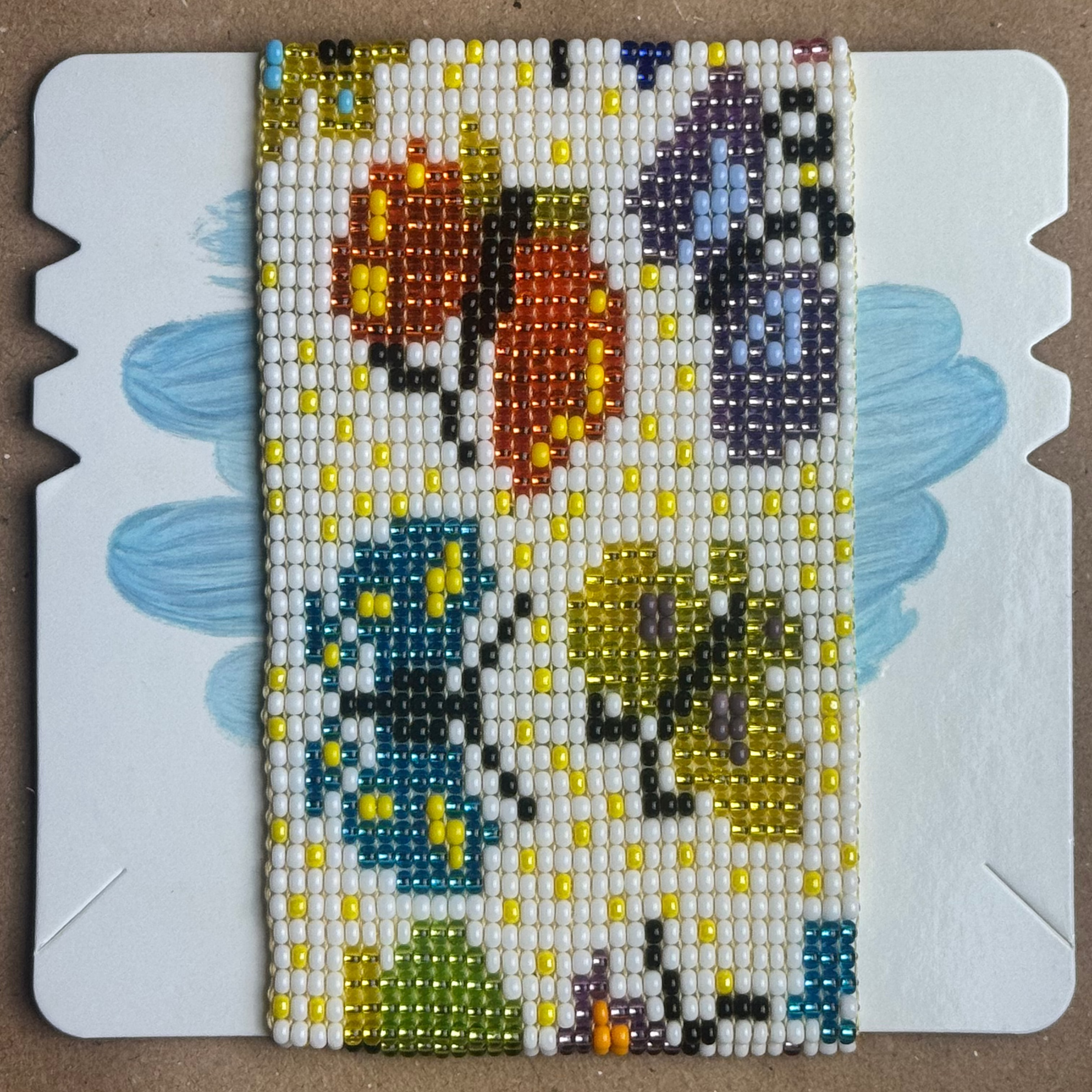 A rectangular beaded canvas featuring a butterfly and floral design, mounted on a white display card with scalloped edges, set against a brown cardboard background.