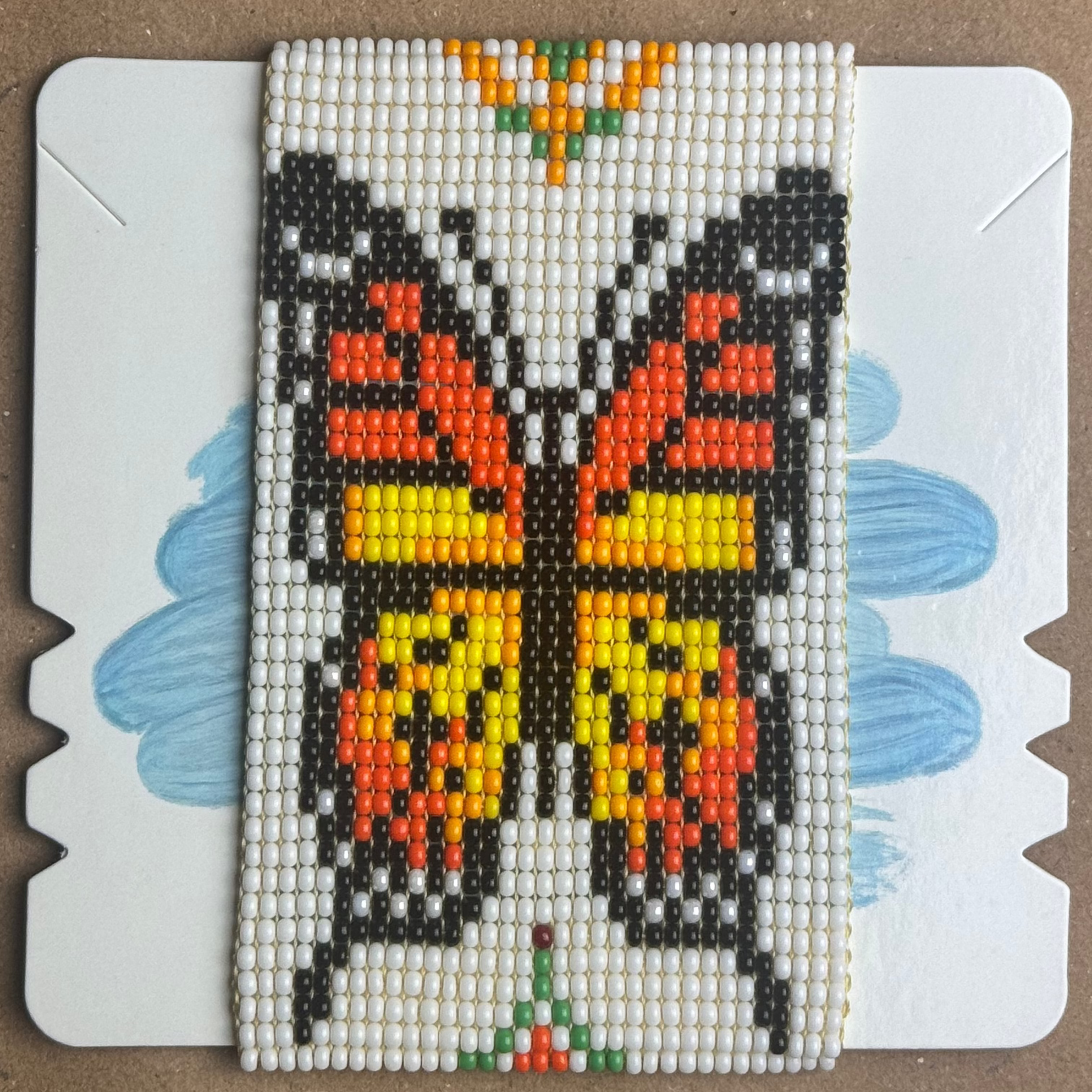 A square beaded mosaic art piece depicting a butterfly, mounted on a white backing card with blue painted wing shapes, placed on a brown cardboard background.